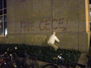 Leslie spray paints "Free CC Now!" in purple and yellow on the concrete walls of the Hennepin County Courthouse.